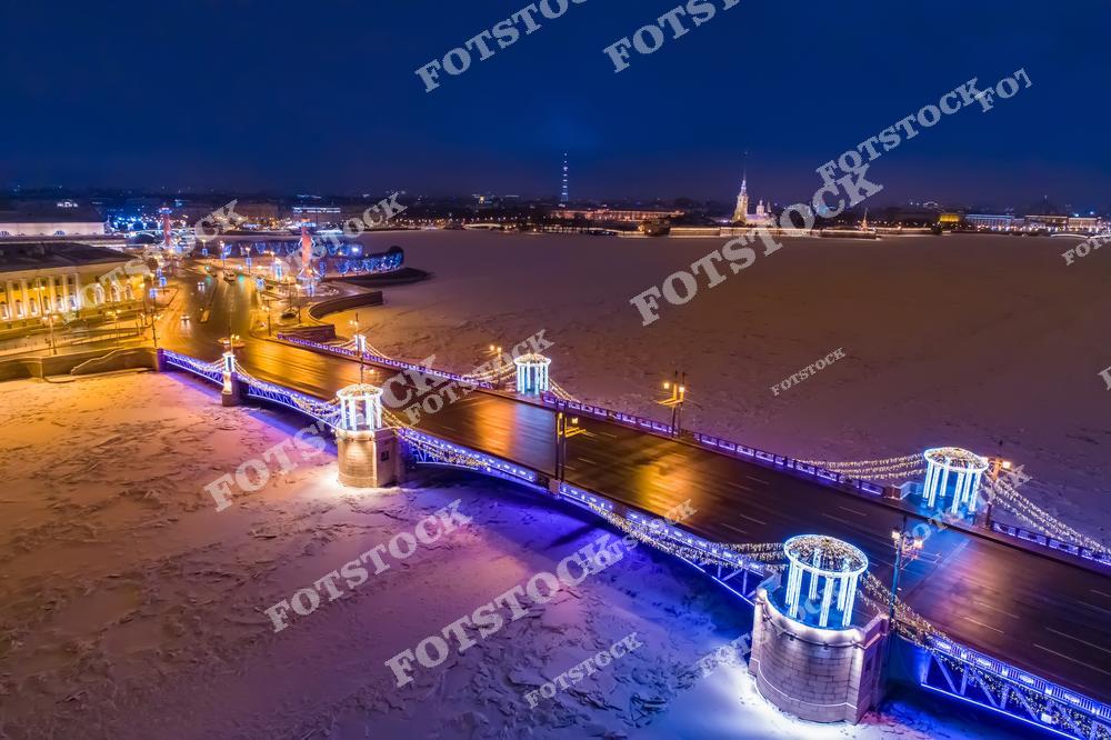 На фотографии изображен Дворцовый мост в ночной подсветке, украшенный гирляндами к зимним праздникам. Под мостом раскинулась замерзшая Нева, покрытая льдом. На заднем плане видна Петропавловская крепость с подсвеченным шпилем. Левее расположено здание Эрмитажа, создающее дополнительный акцент на красоте зимнего Петербурга. Ночная иллюминация подчеркивает торжественный облик города.